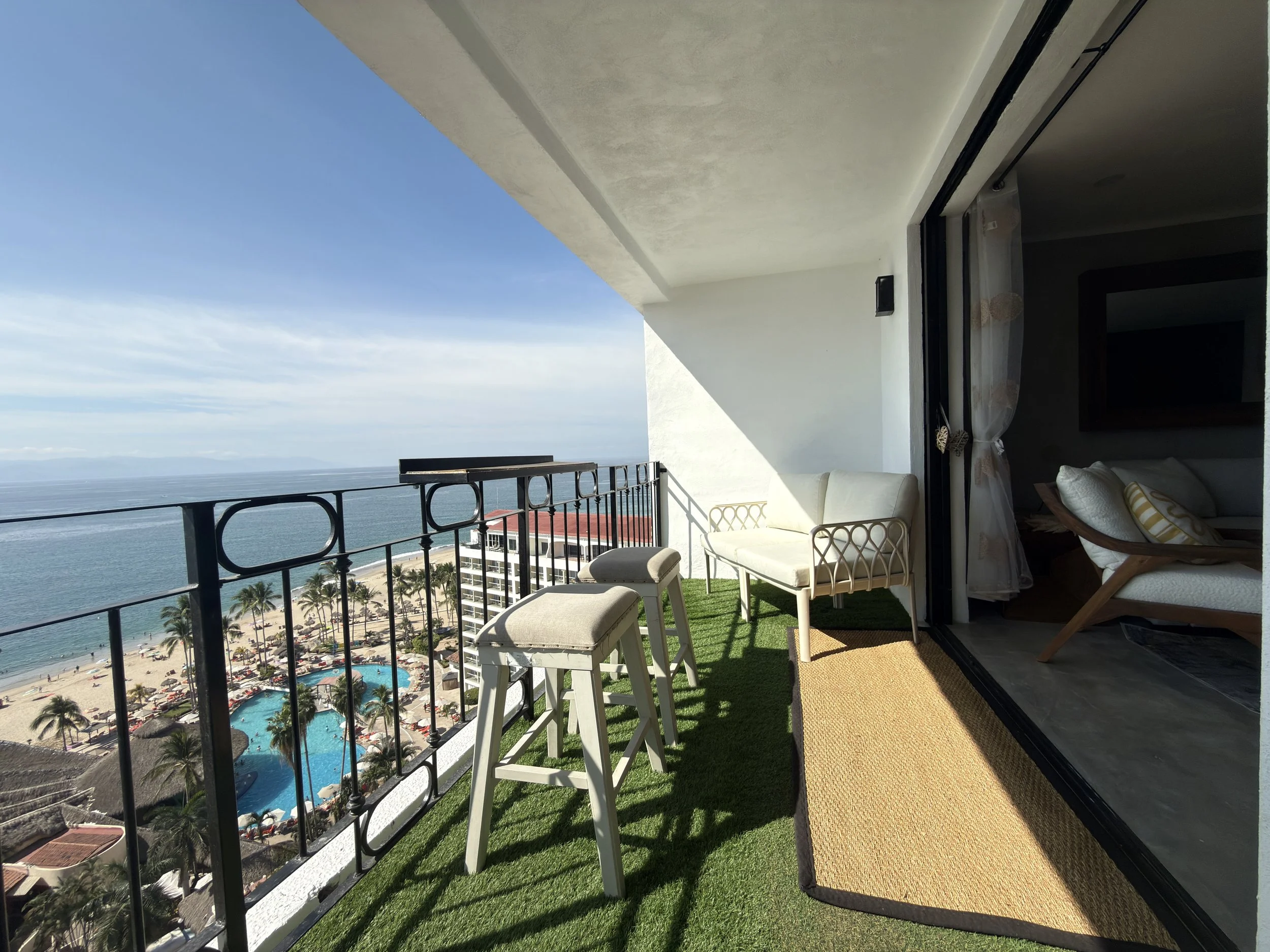 Balcony with chairs and a view of a beach, palm trees, and pools.