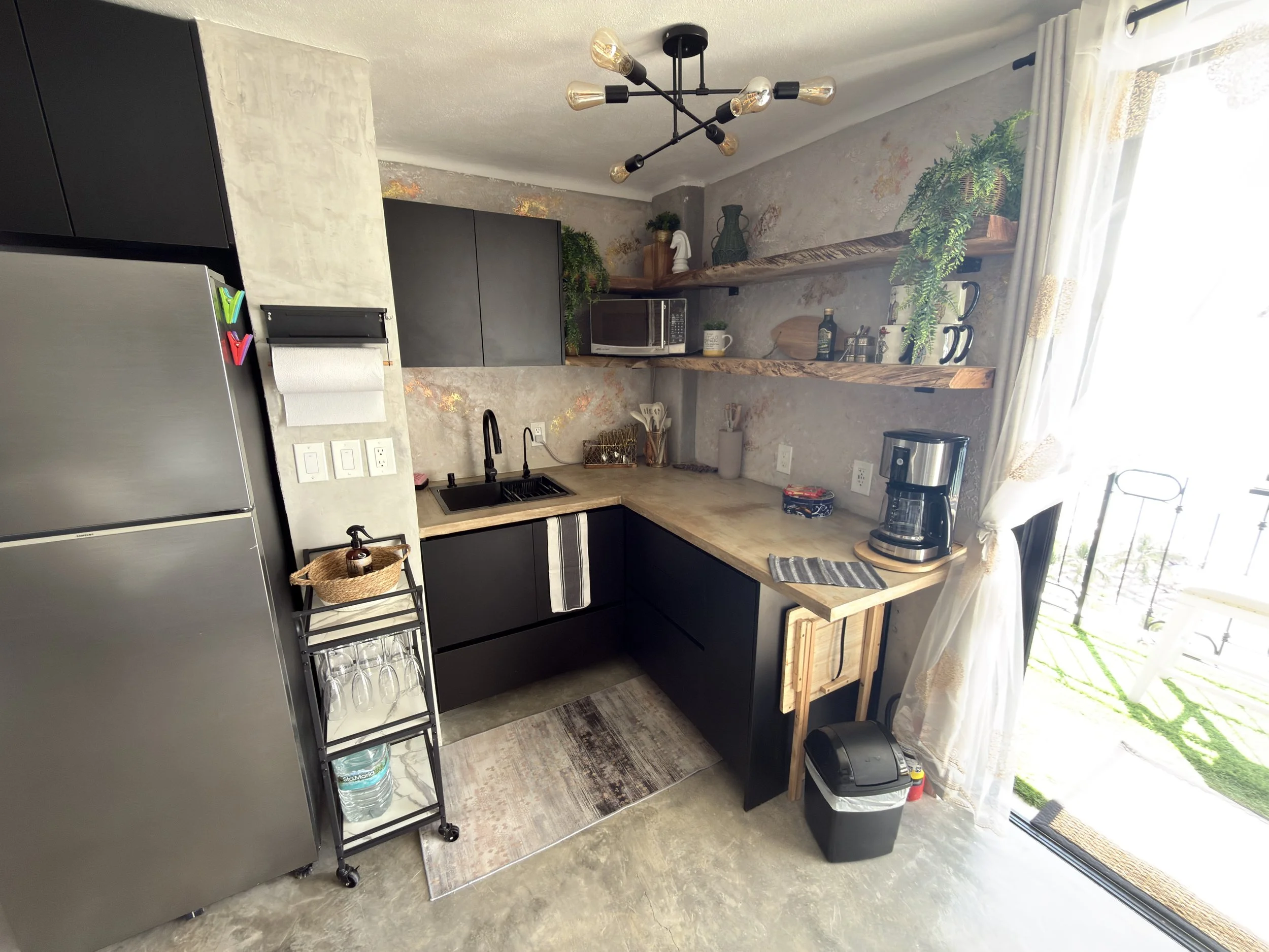 Modern kitchen with black cabinets, stainless steel refrigerator, open wooden shelves with plants and kitchenware, small black trash bin, coffee maker, microwave, and large window with white curtains.
