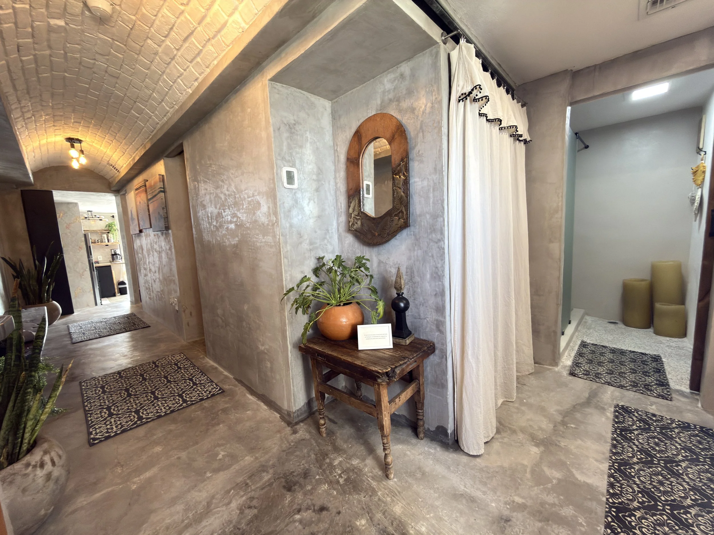 Interior of a modern home with concrete walls and flooring, featuring a hallway with patterned rugs, a small wooden table with a plant and decorative items, and a curtain-covered shower area with beige stools nearby.