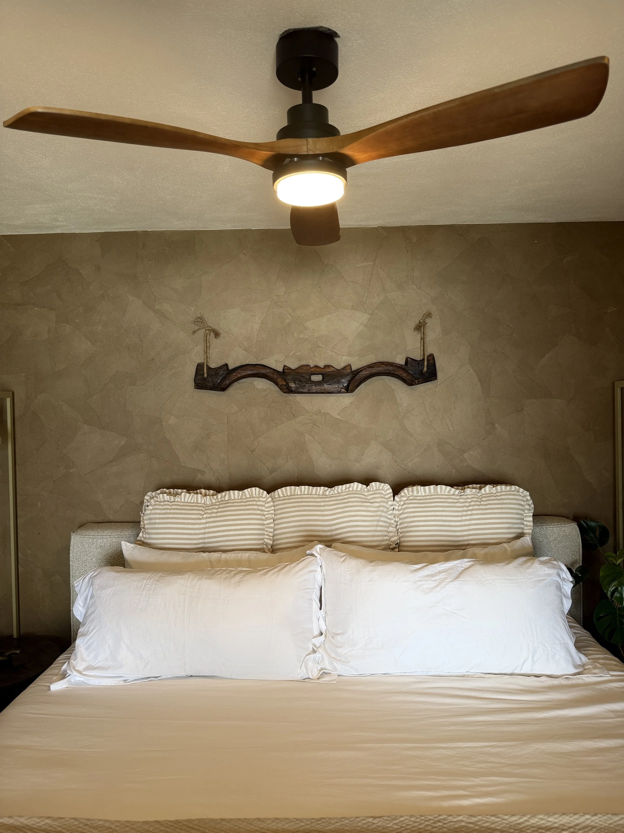 A bedroom with a bed featuring white sheets and pillows, a textured beige headboard, four striped pillows, a wooden wall art piece above the bed, and a ceiling fan with wooden blades and a light fixture.