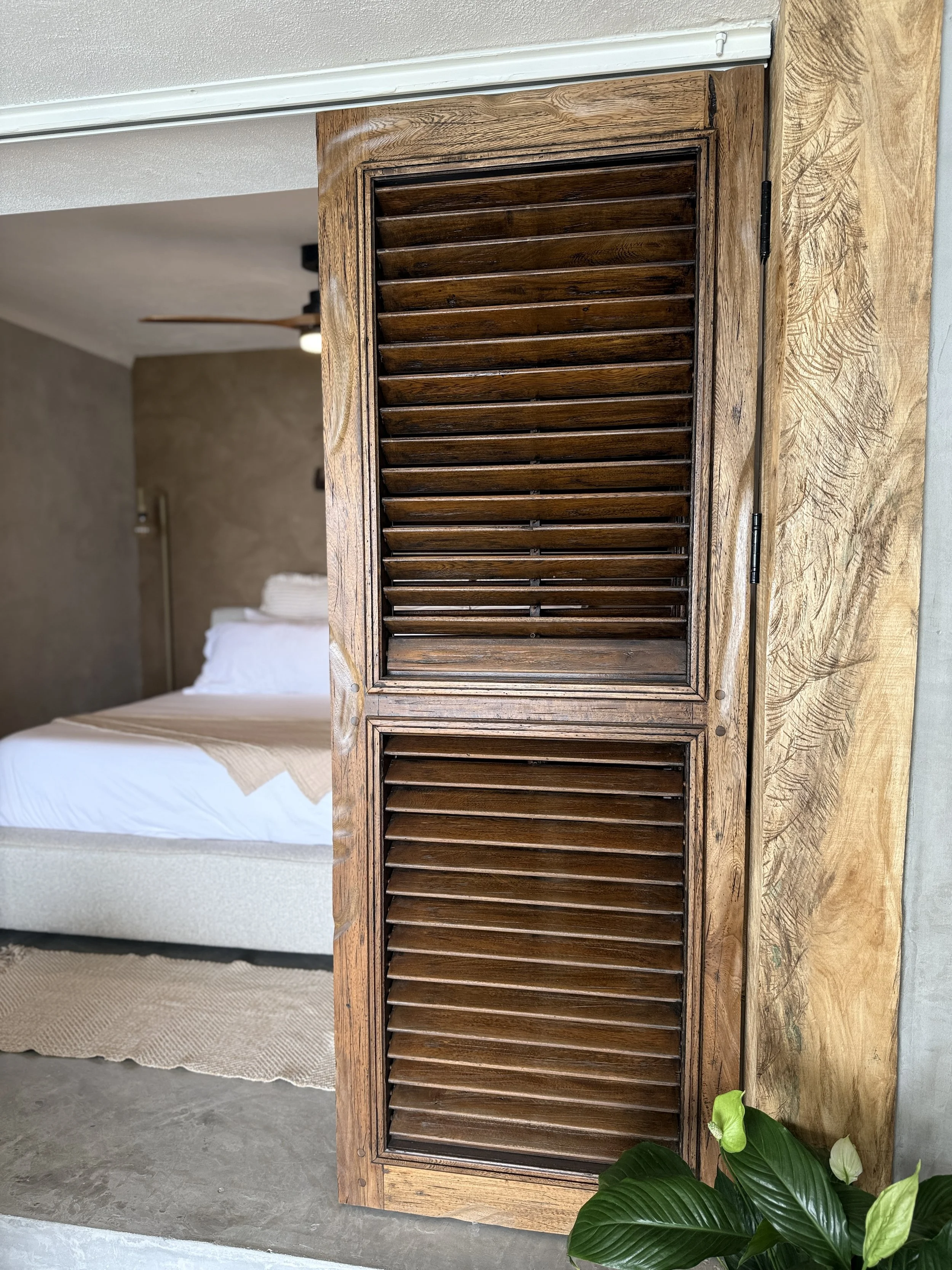 A wooden louvered door or cabinet on a wall next to a bedroom with a bed, beige walls, and a ceiling fan with wooden blades.