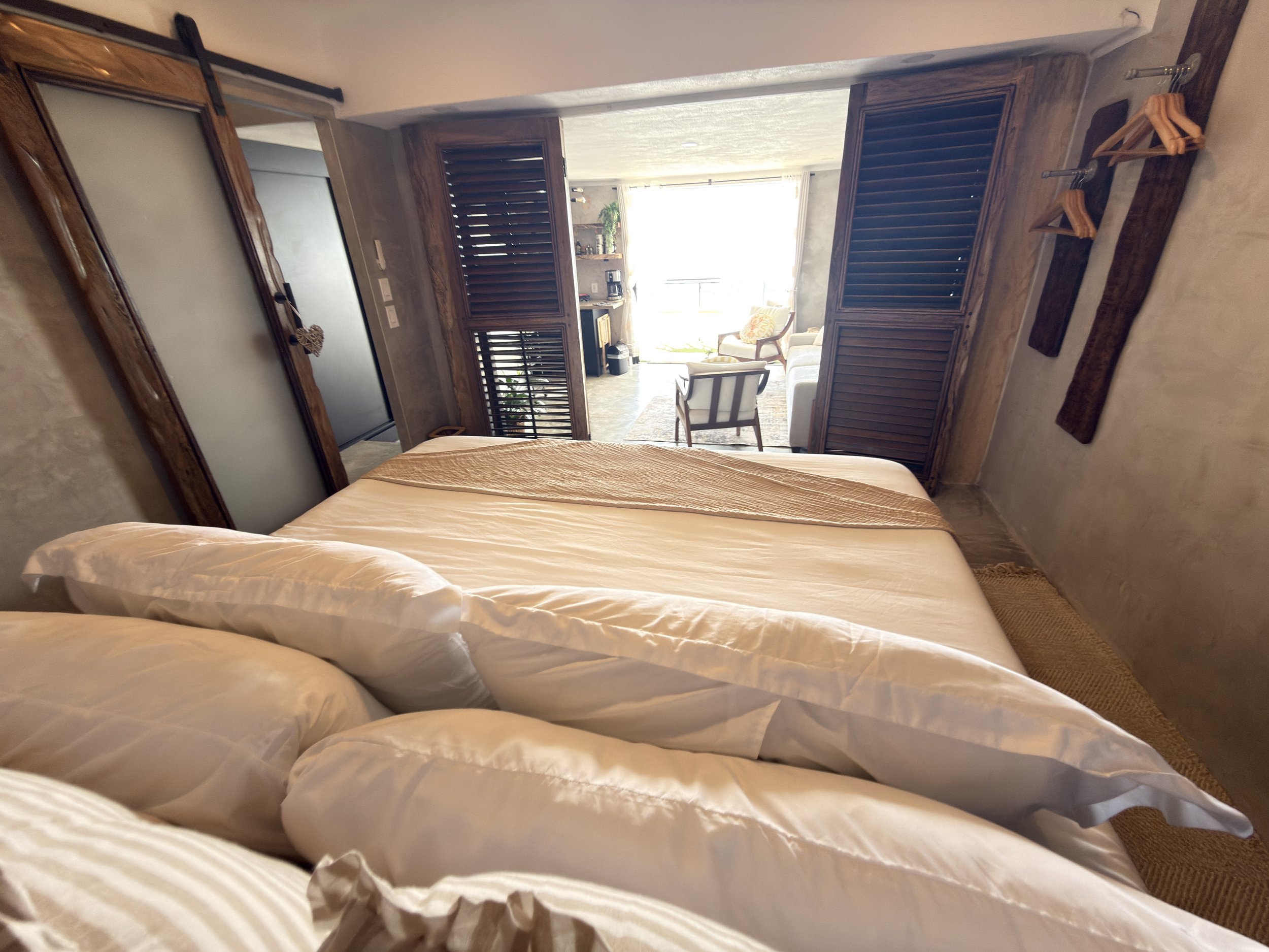 A bedroom with a bed in the foreground and an adjacent living area visible through wooden shutters, with chairs and a balcony outside.
