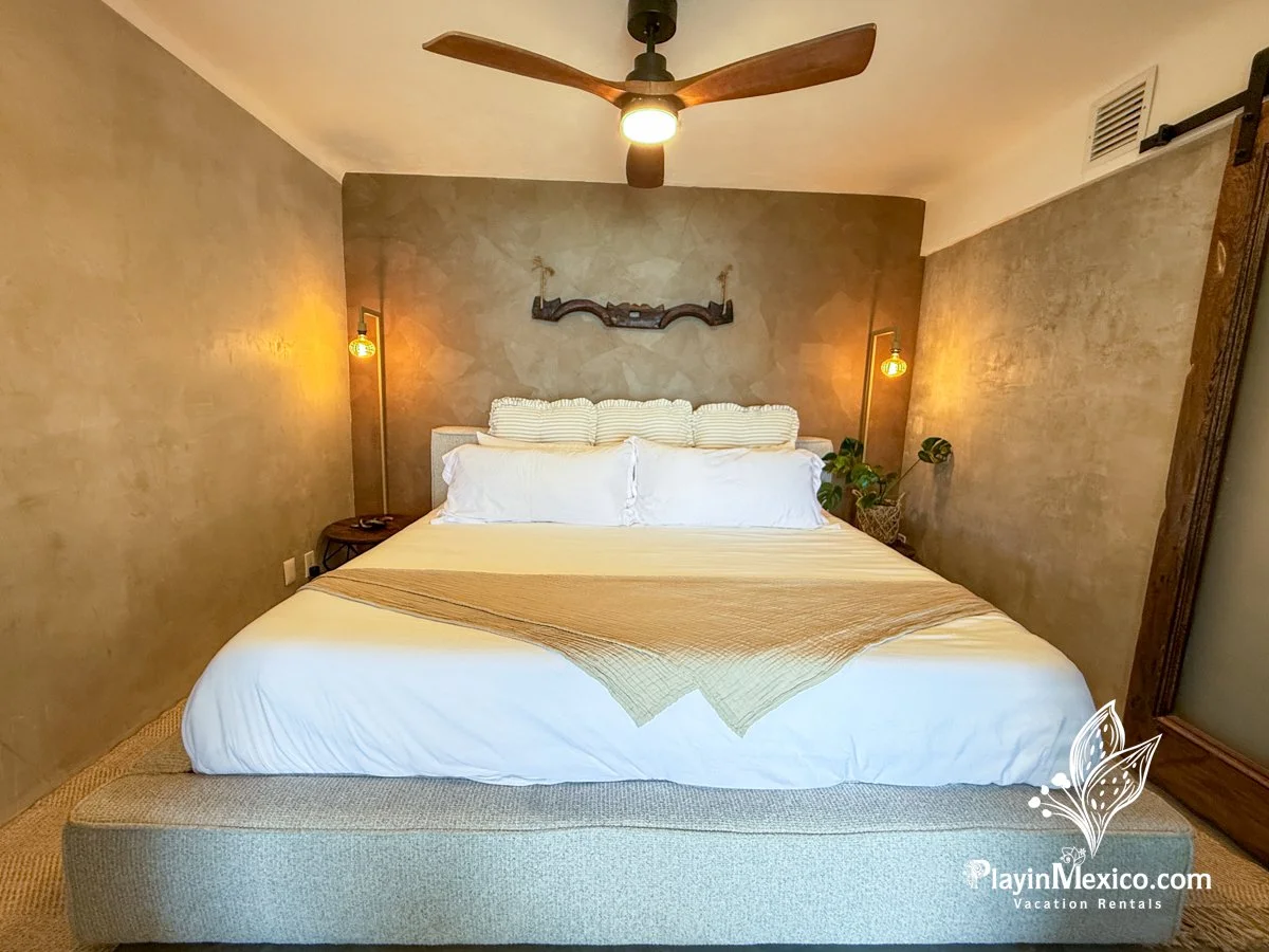 A neatly made bed with white linens and striped pillows, flanked by two bedside lamps, in a cozy bedroom with a ceiling fan and wall decoration.