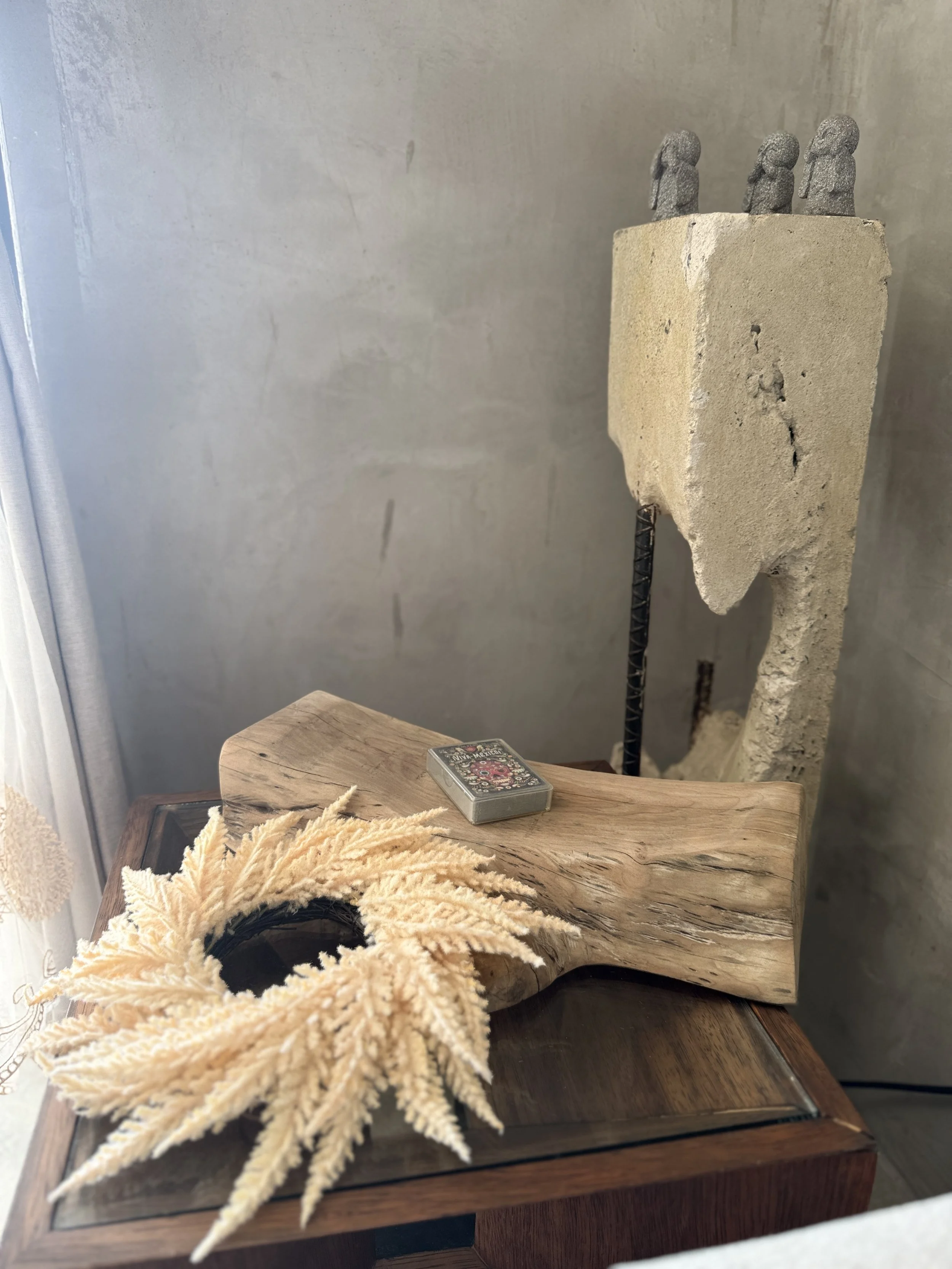 A wooden table with a deck of playing cards, a decorative beige feather arrangement, and a carved wooden object. In the background, a concrete sculpture with three head-shaped figures on top.