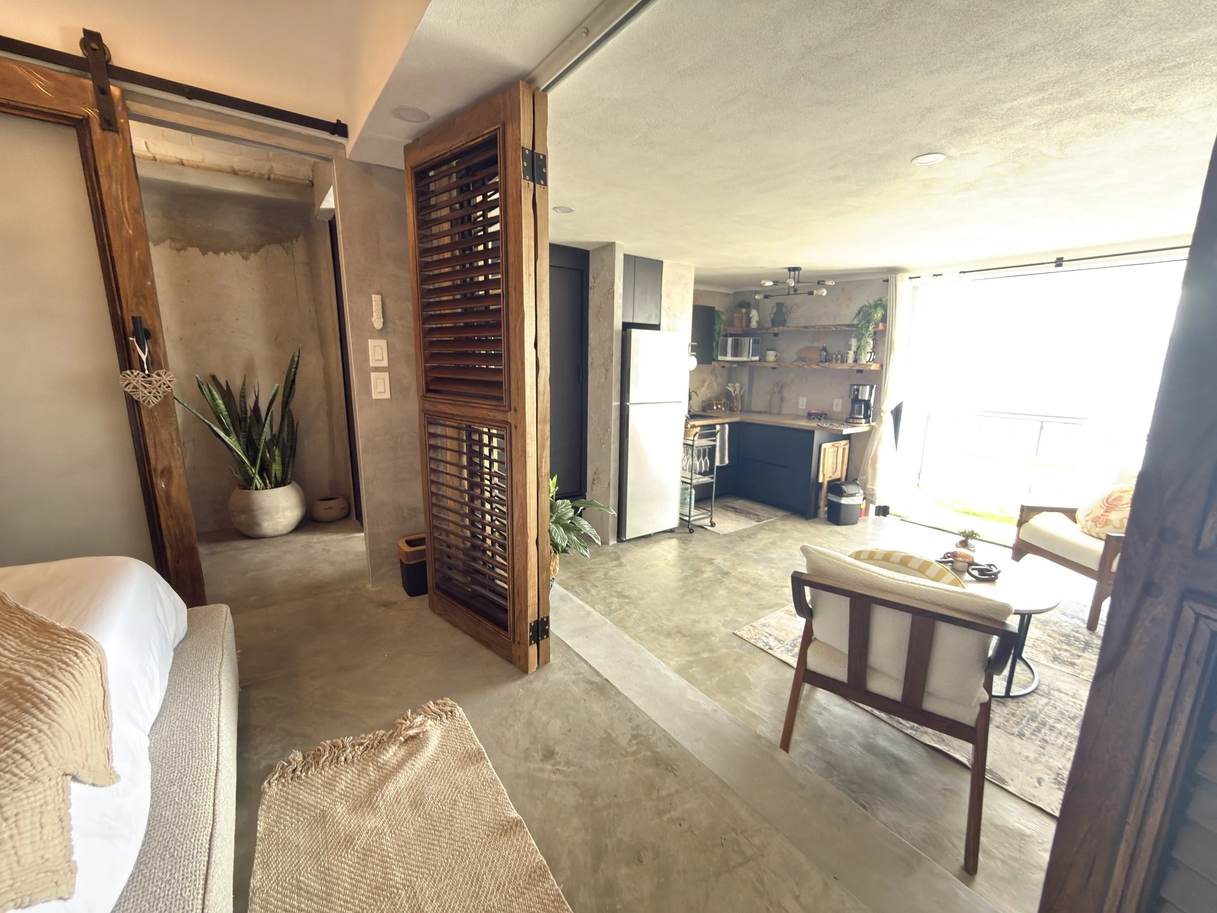Open-concept living space with kitchen, seating area, and large window with sunlight. Wood accents and plants decorate the room.