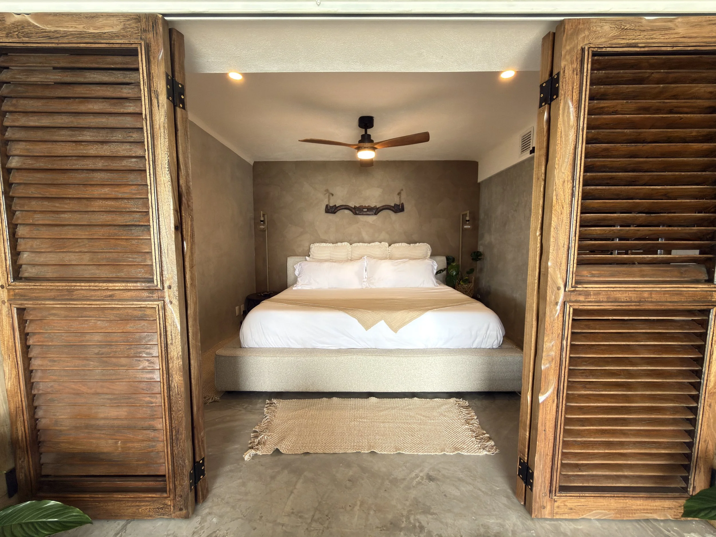 A bedroom with a bed and wooden shutters framing the view, beige walls, and a ceiling fan.