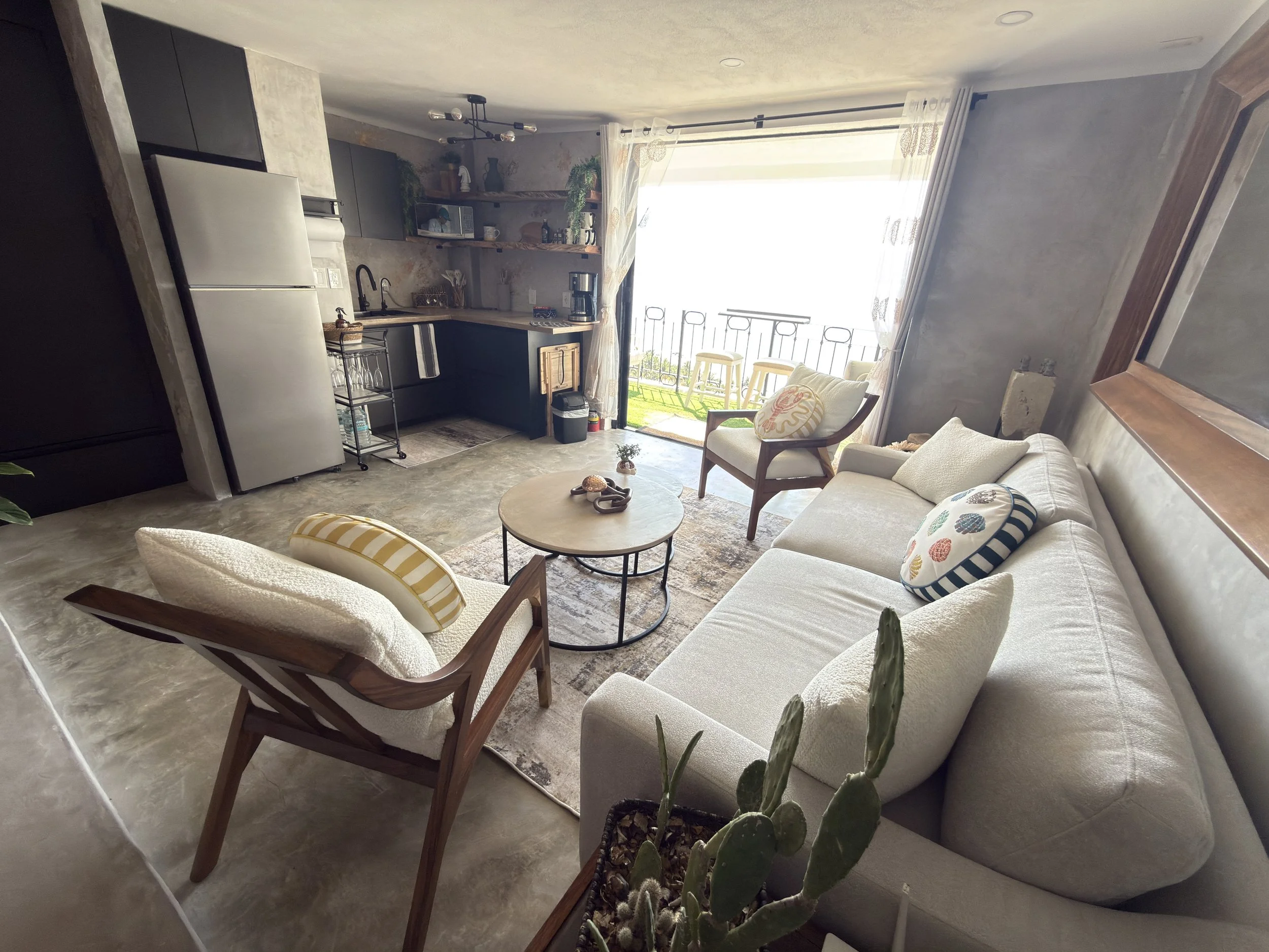 Living room with a white sofa, wooden armchair, coffee table, and a small balcony with outdoor seating in the background.