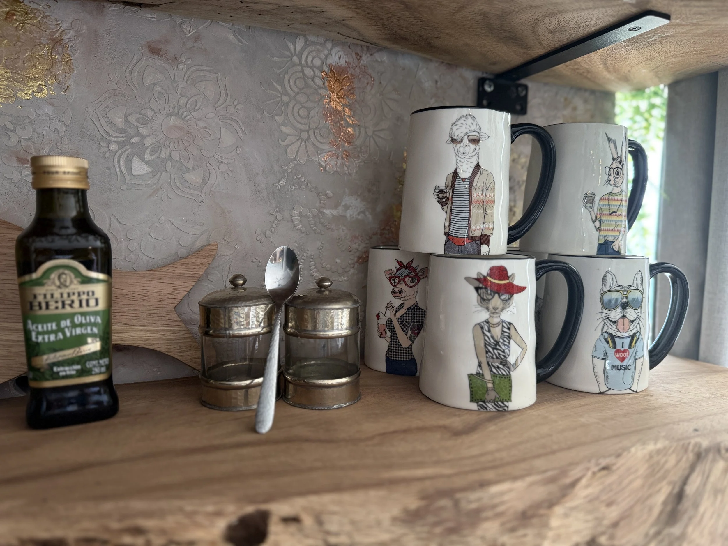 A kitchen shelf holds a small bottle of extra virgin olive oil, two glass spice containers with silver lids, a silver spoon, and four coffee mugs with illustrations of cats dressed fashionably.