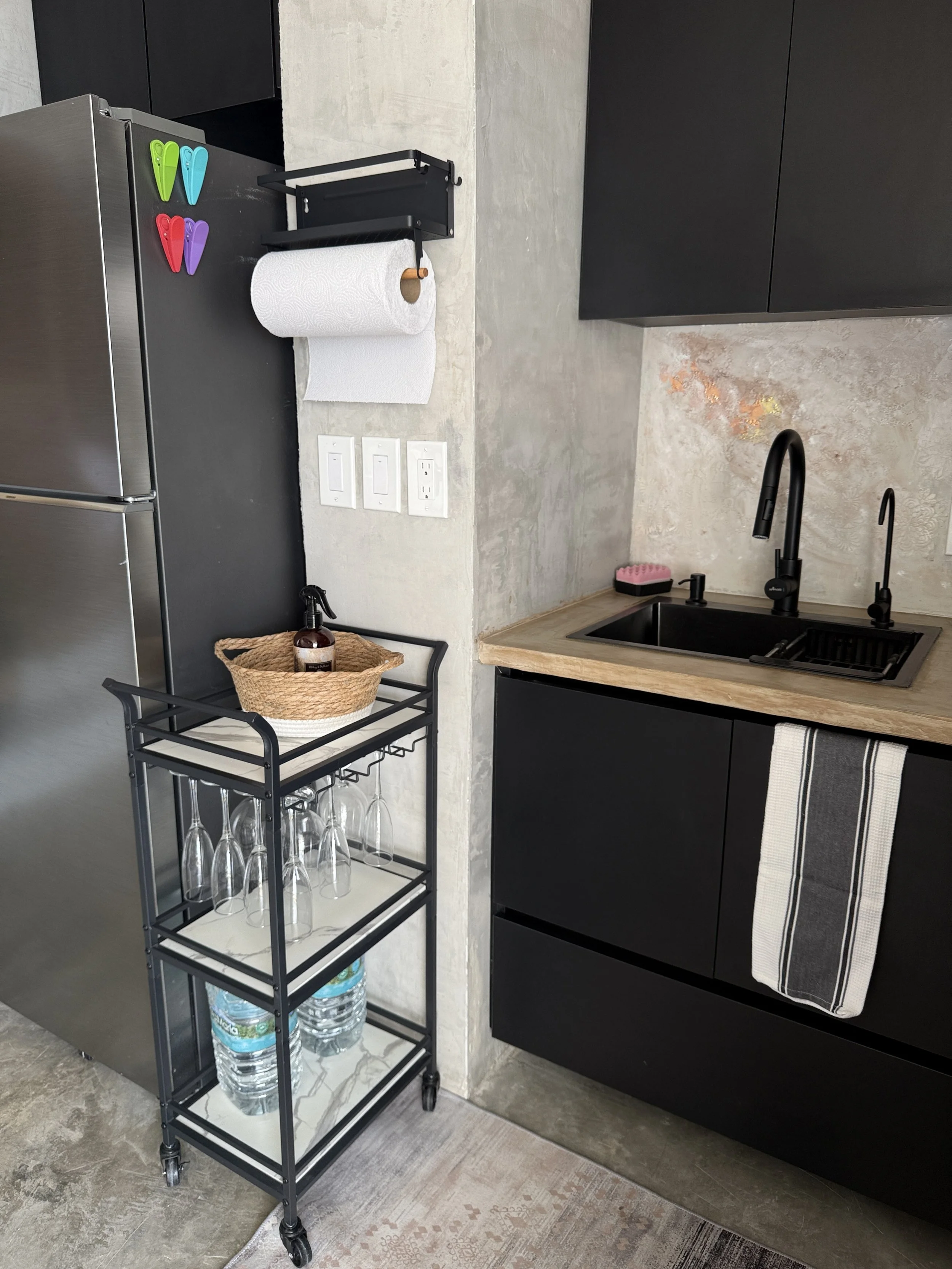 A modern kitchen corner with a black metal cart holding glasses and bottled water, a wine bottle in a basket, a black faucet and sink, dark cabinets, a paper towel roll, and colorful clips on the fridge.