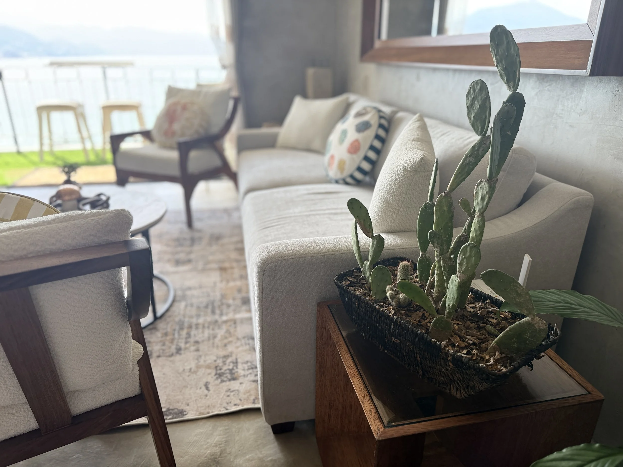 Interior of a cozy living room with a beige sofa, colorful pillows, a wooden side table with a potted cactus, and a balcony with chairs overlooking a body of water and mountains.