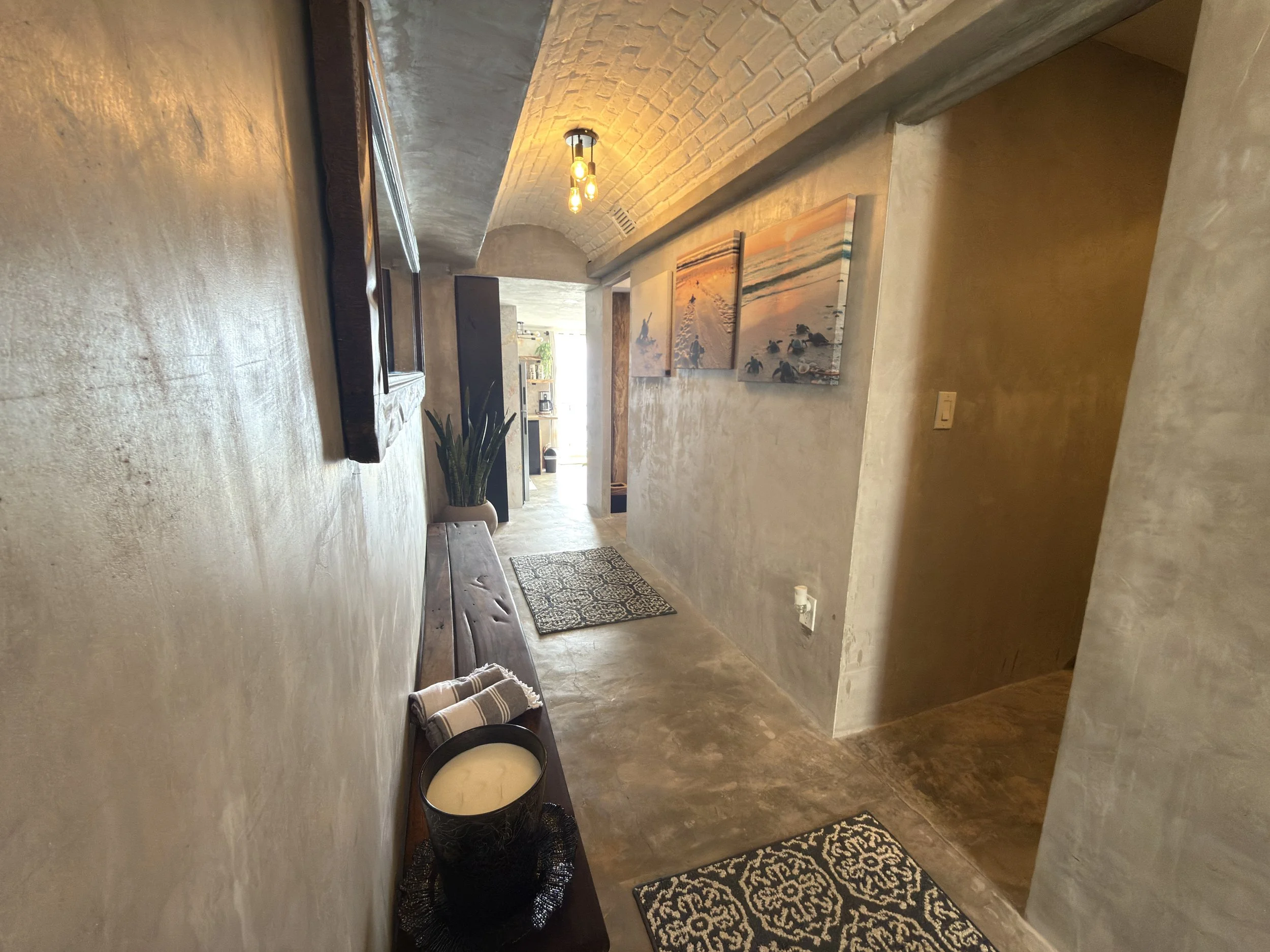 A hallway with cream-colored textured walls, a brick vaulted ceiling, and a wooden bench with decor items including a candle holder and towels. Wall art with sunset and ocean scenes, a potted plant, and light fixtures on the ceiling are visible.