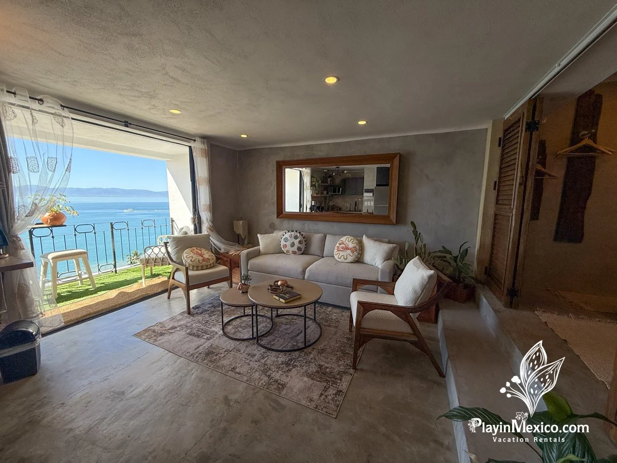Living room with large glass sliding door opening to a balcony with ocean view, furnished with a white sofa, armchairs, round coffee tables, decorative pillows, and indoor plants, with kitchen visible through a window mirror.