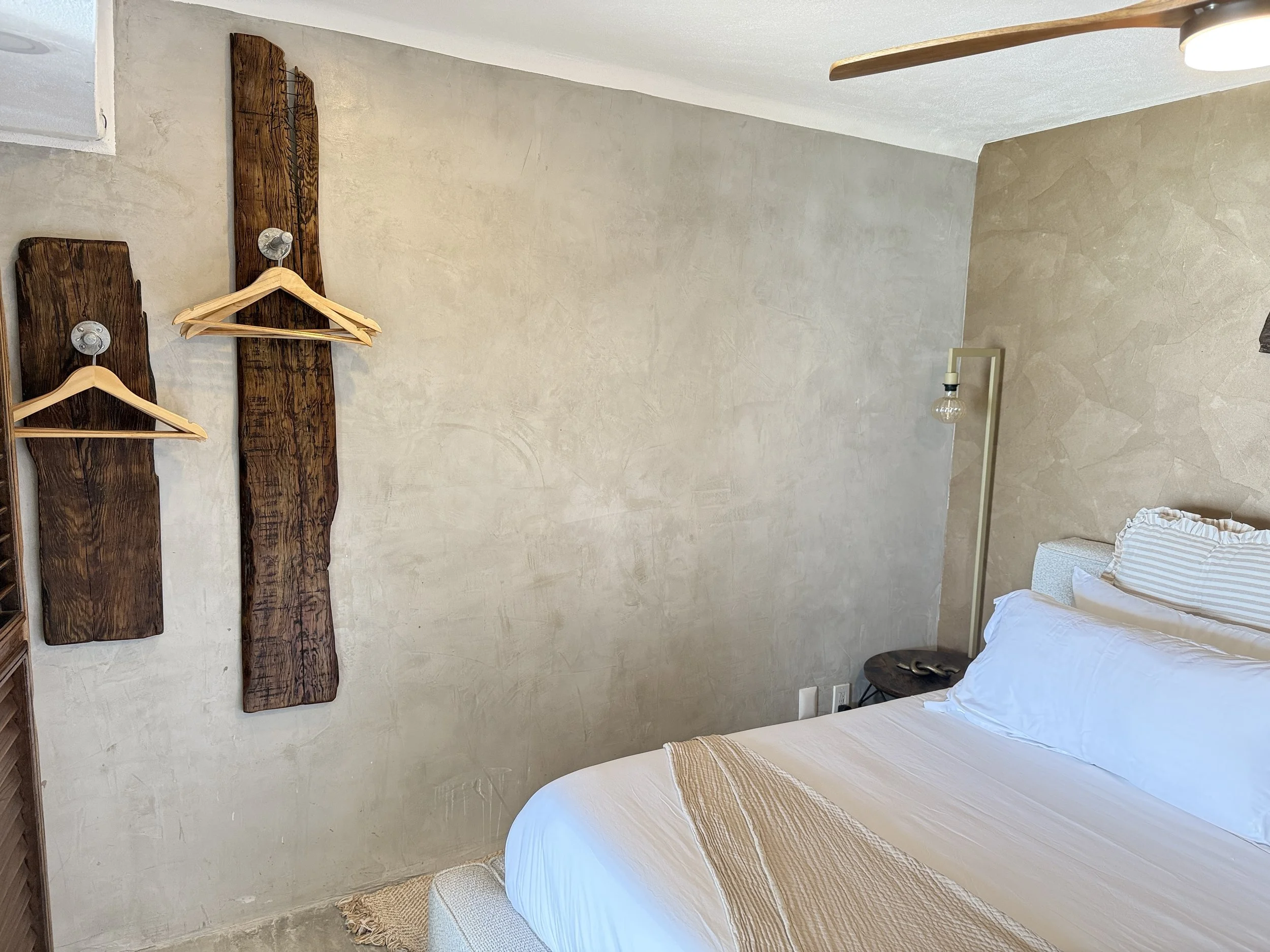 A minimalist bedroom with a bed dressed in white linens, a black nightstand, a tan textured wall, a wooden and string lamp, and a rustic wooden wall decor with hangers.