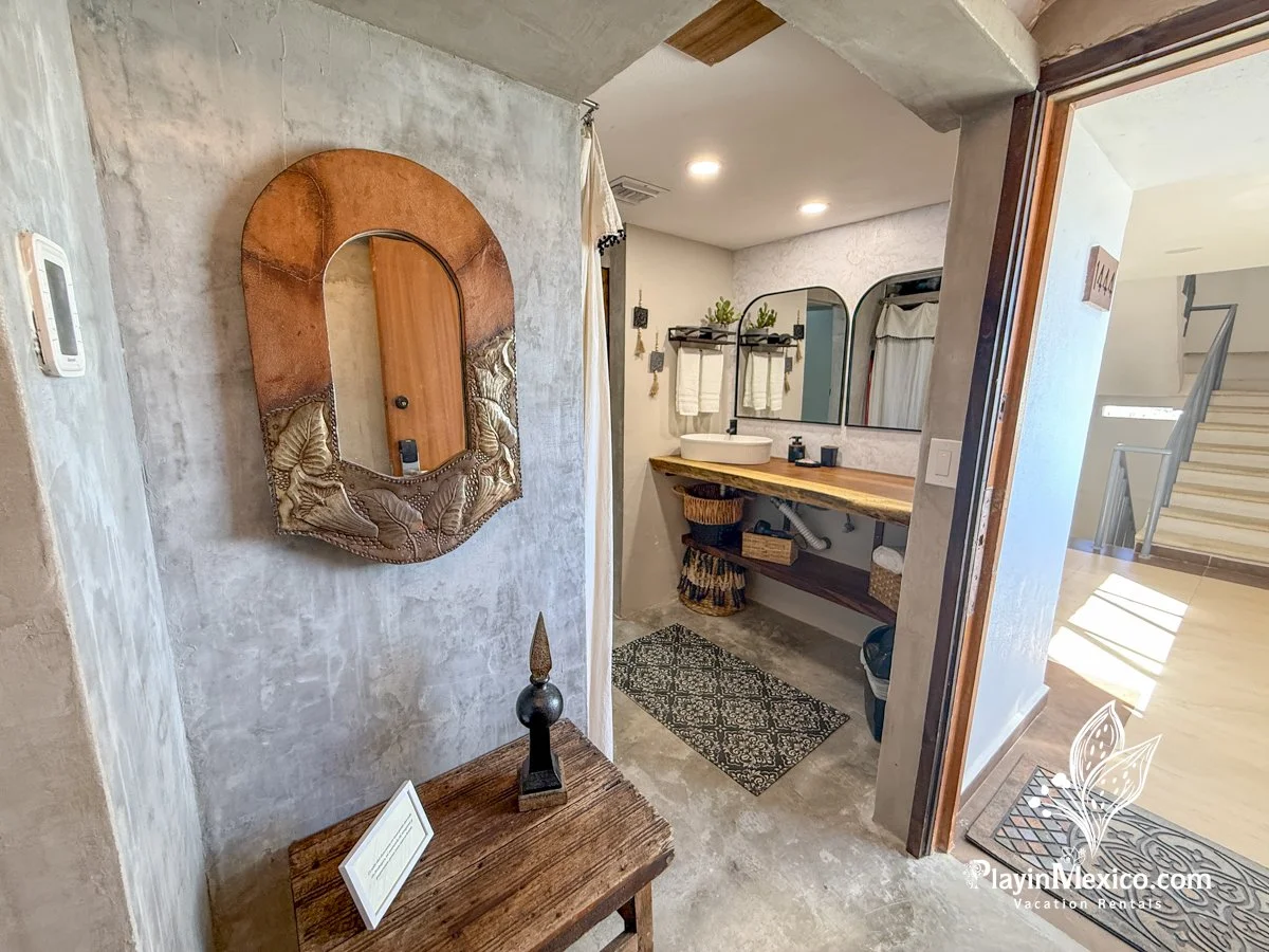 Bathroom with a wooden countertop, sinks, and a large mirror, next to an abstract mirror with a metallic frame, and a staircase in the background.