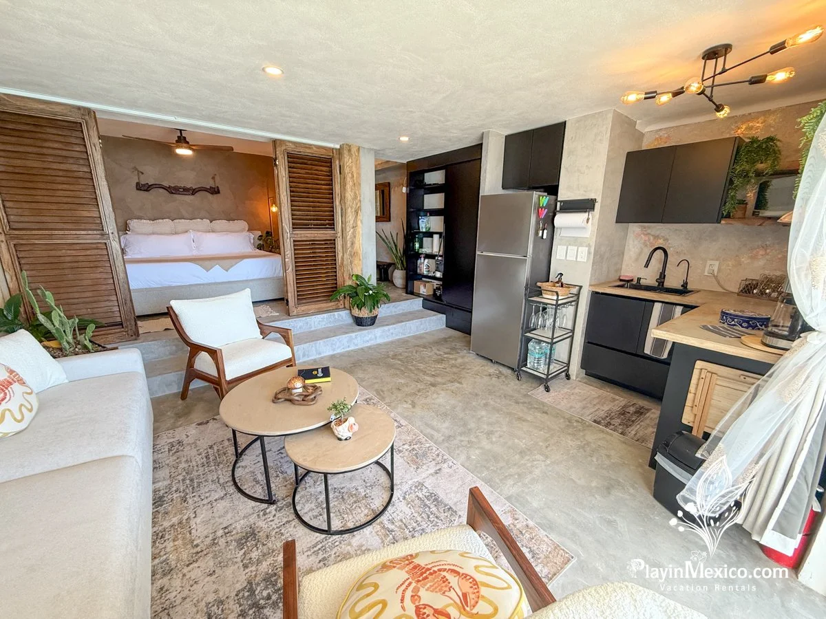 Open-concept living area with a sitting space and kitchen, bedroom separated by wooden shutters, modern decor, and plants.
