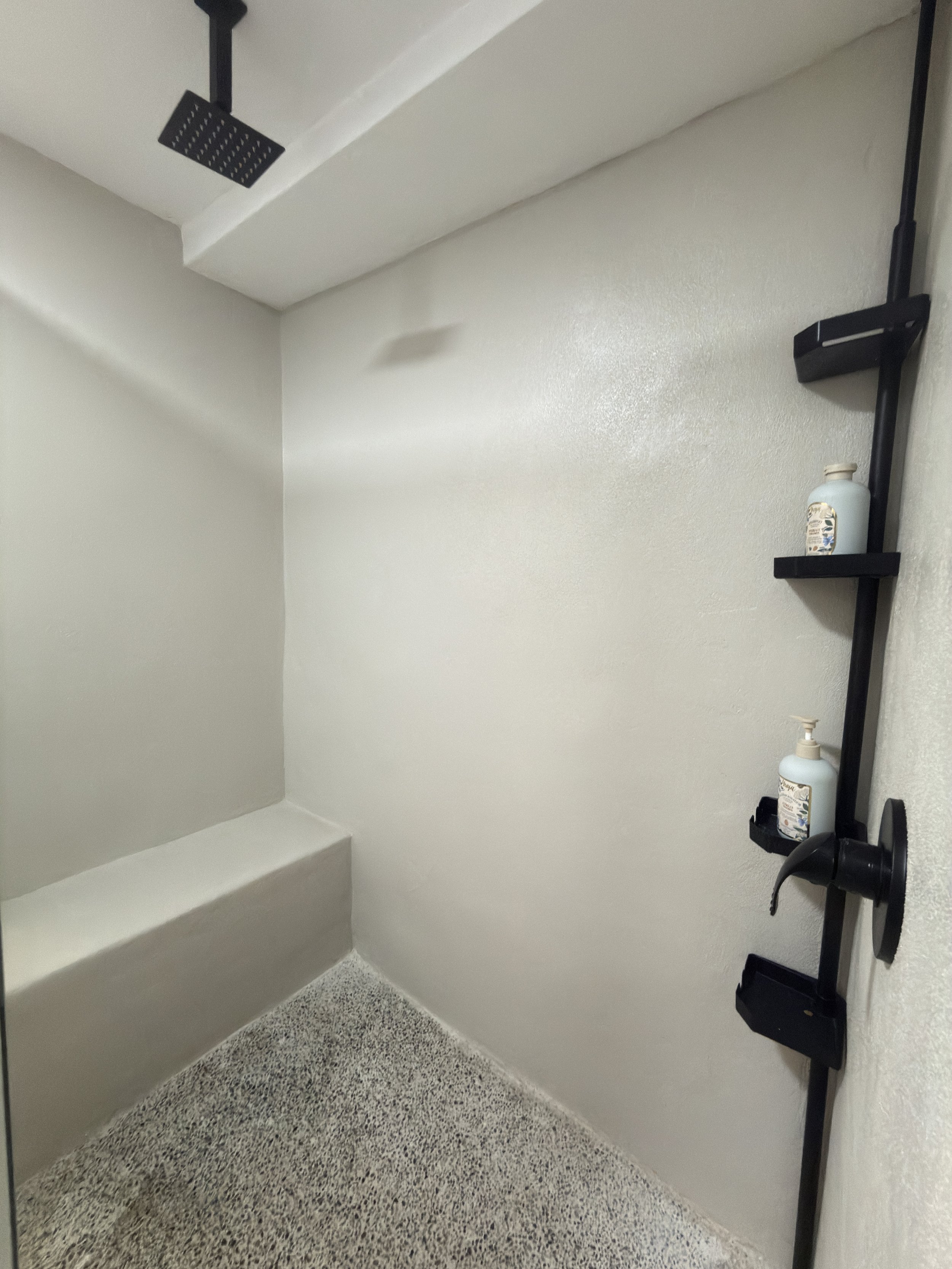 Empty shower with a black rainfall showerhead, built-in bench, and a black wall-mounted soap and shampoo holder with two bottles.