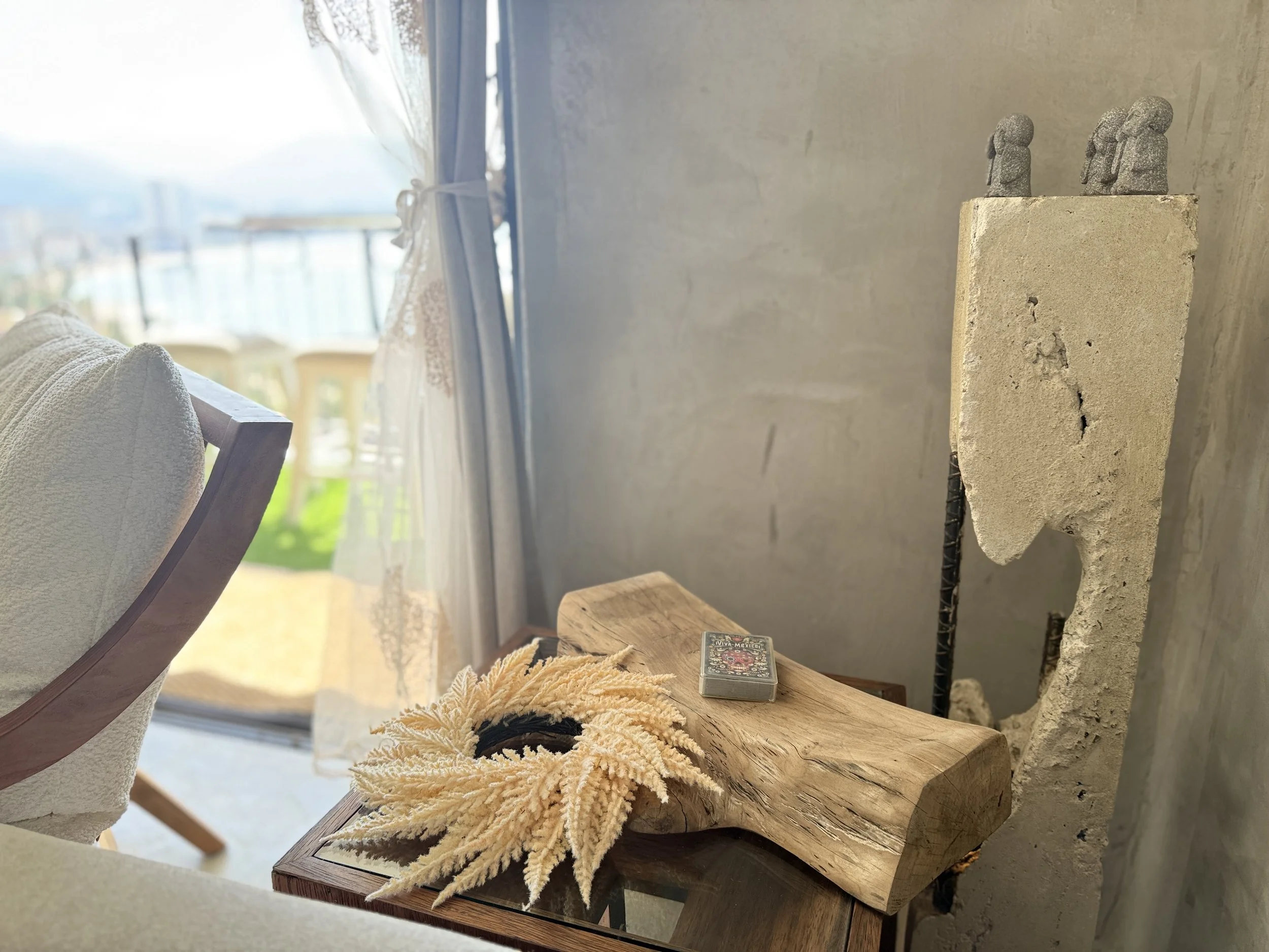 Interior view of a cozy room with a window revealing a balcony and cityscape, a wooden table holding a deck of cards and a decorative dried flower arrangement, a gray upholstered chair, and a tall textured sculpture on the wall.