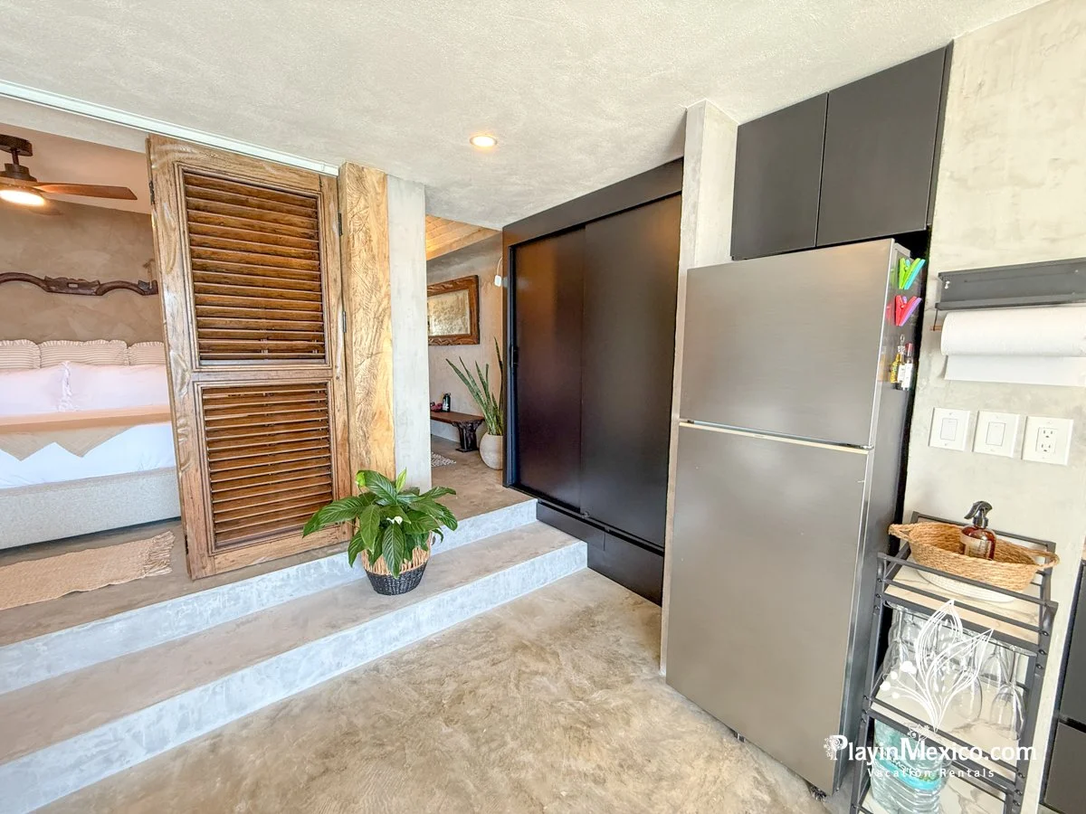 Kitchen area with a stainless steel refrigerator, black upper cabinets, a small shelving cart, and a glimpse of a bedroom with a bed and wooden headboard in the background.