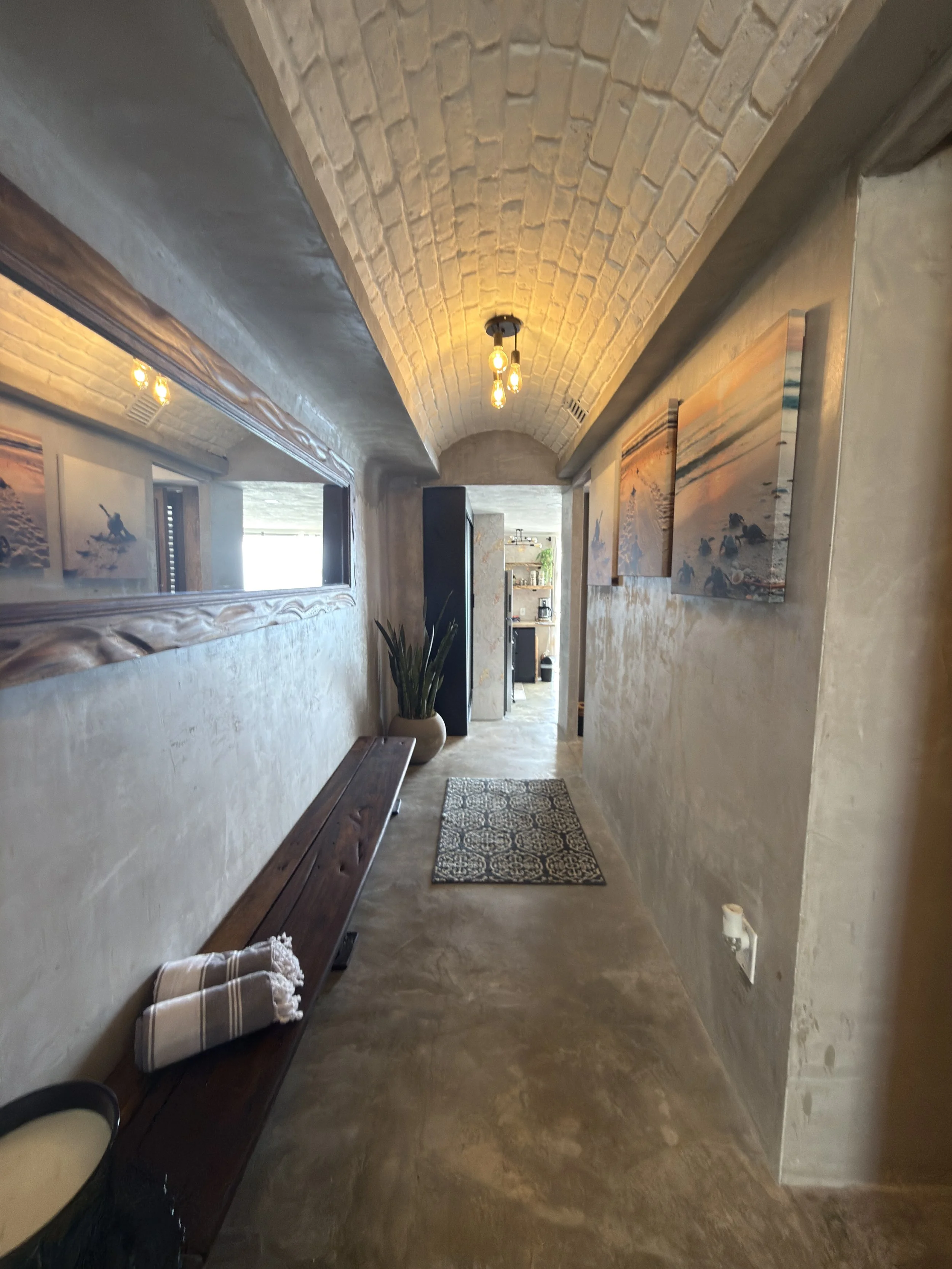 A hallway with a vaulted brick ceiling, wall-mounted artwork, a wooden bench, potted plant, and patterned rug.