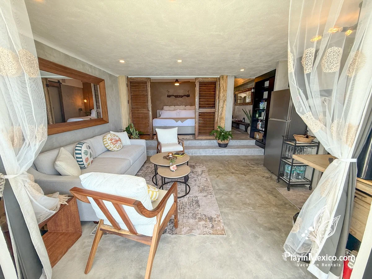 A cozy living room with a beige sofa, wooden armchair, and round coffee tables. There are decorative pillows on the sofa and a large mirror on the wall. The room opens to a bedroom with a bed and louvered wooden doors, and there are houseplants and a