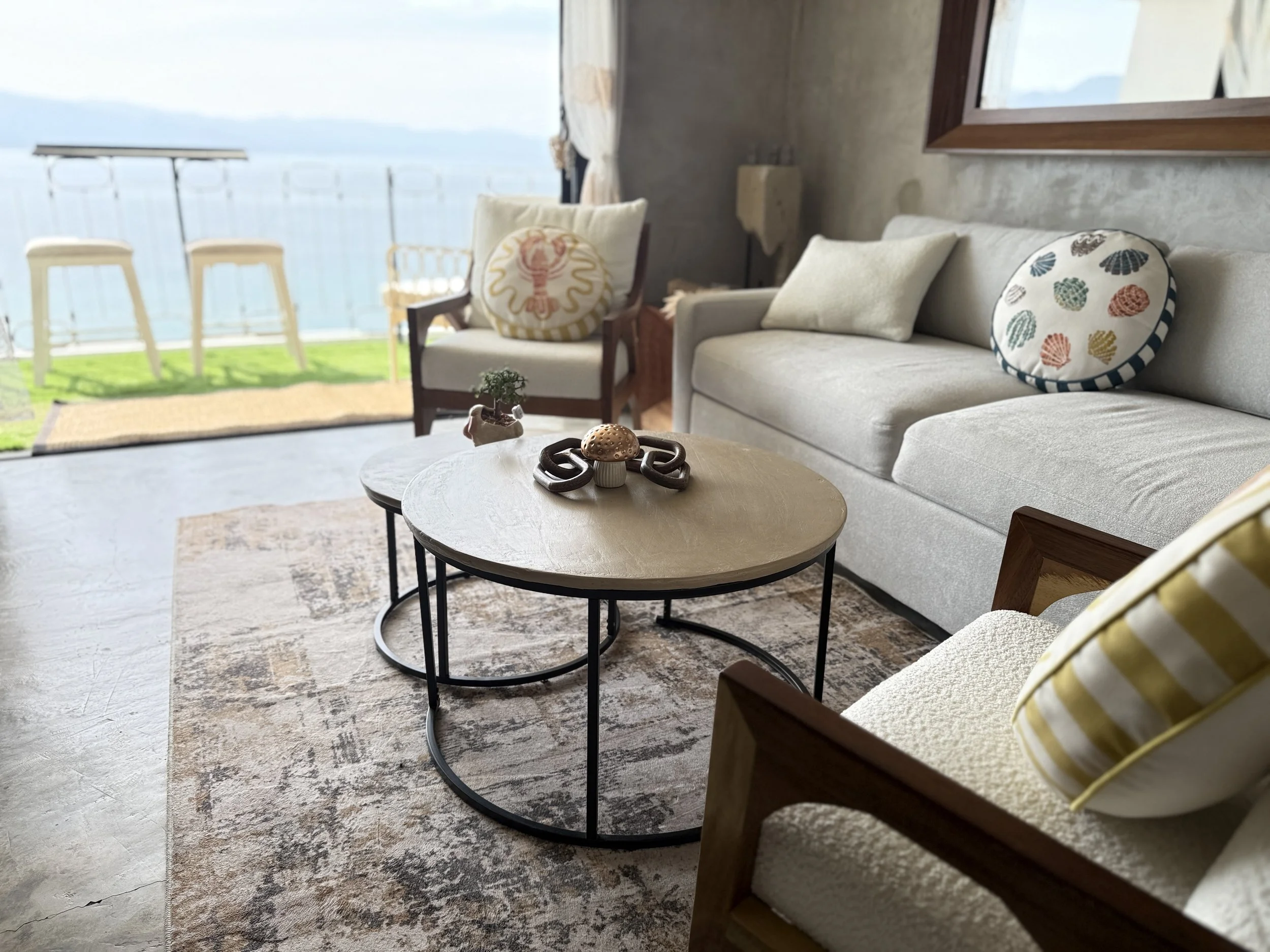 Living room with sofa, armchair, tables, and decorative pillows, overlooking a patio and water view.