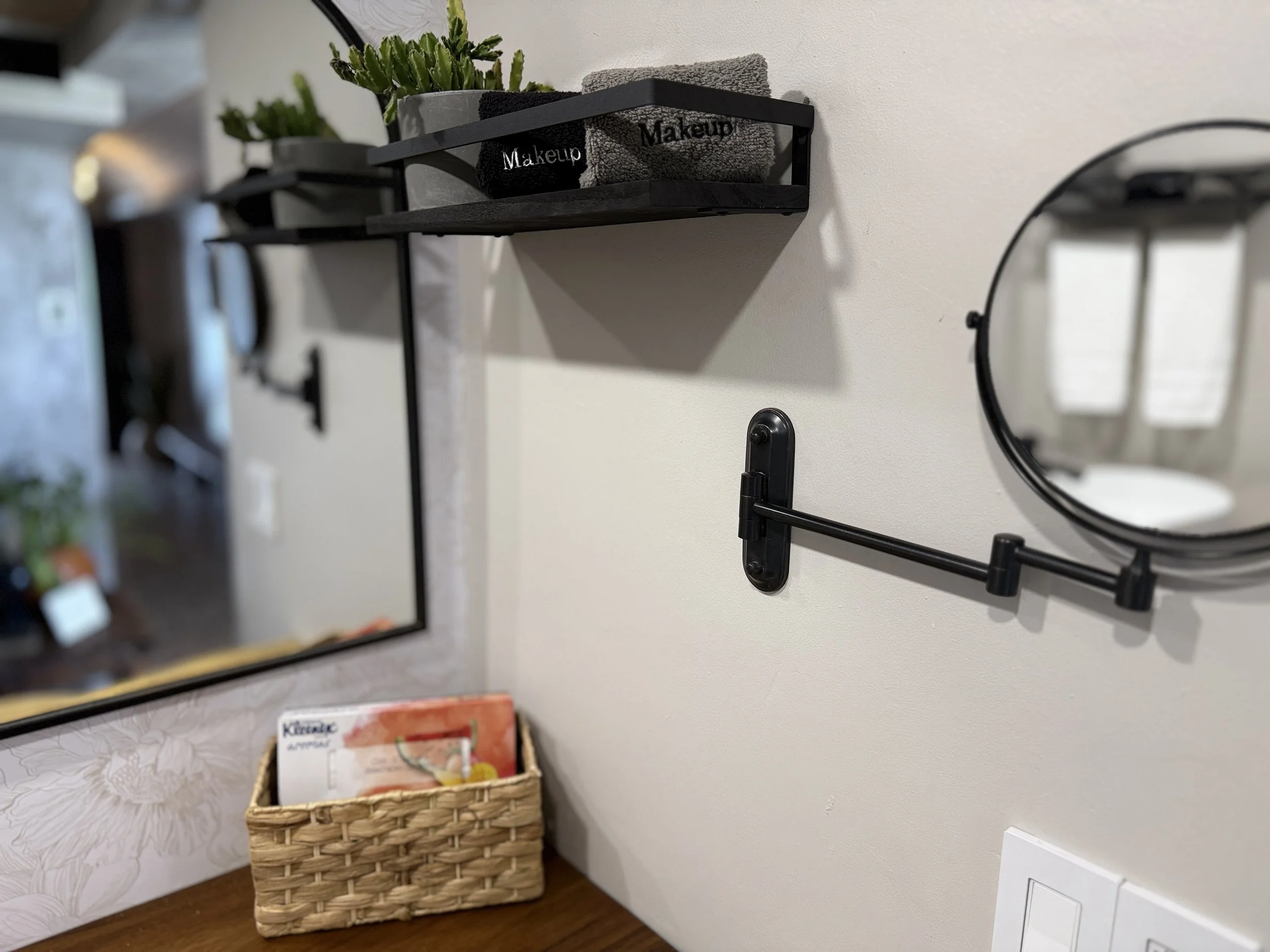 Decorative bathroom wall with a small round mirror, a black wall-mounted towel bar, a black shelf holding small potted plants and towels labeled 'Makeup', and a wicker basket with booklets on a wooden surface.