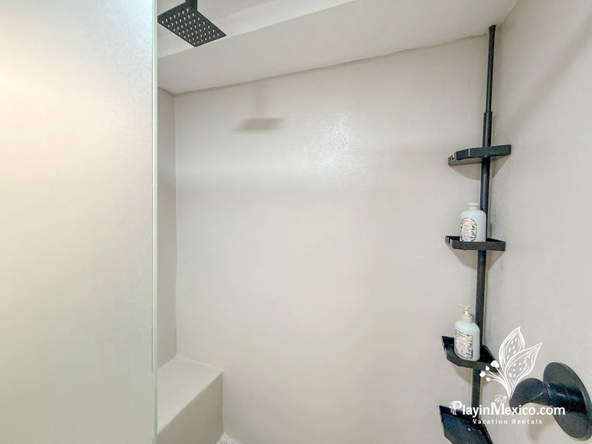 Empty shower with a black wall-mounted shelf holding two bottles of shower gel or shampoo, a black showerhead, and a ventilation fan in the ceiling.