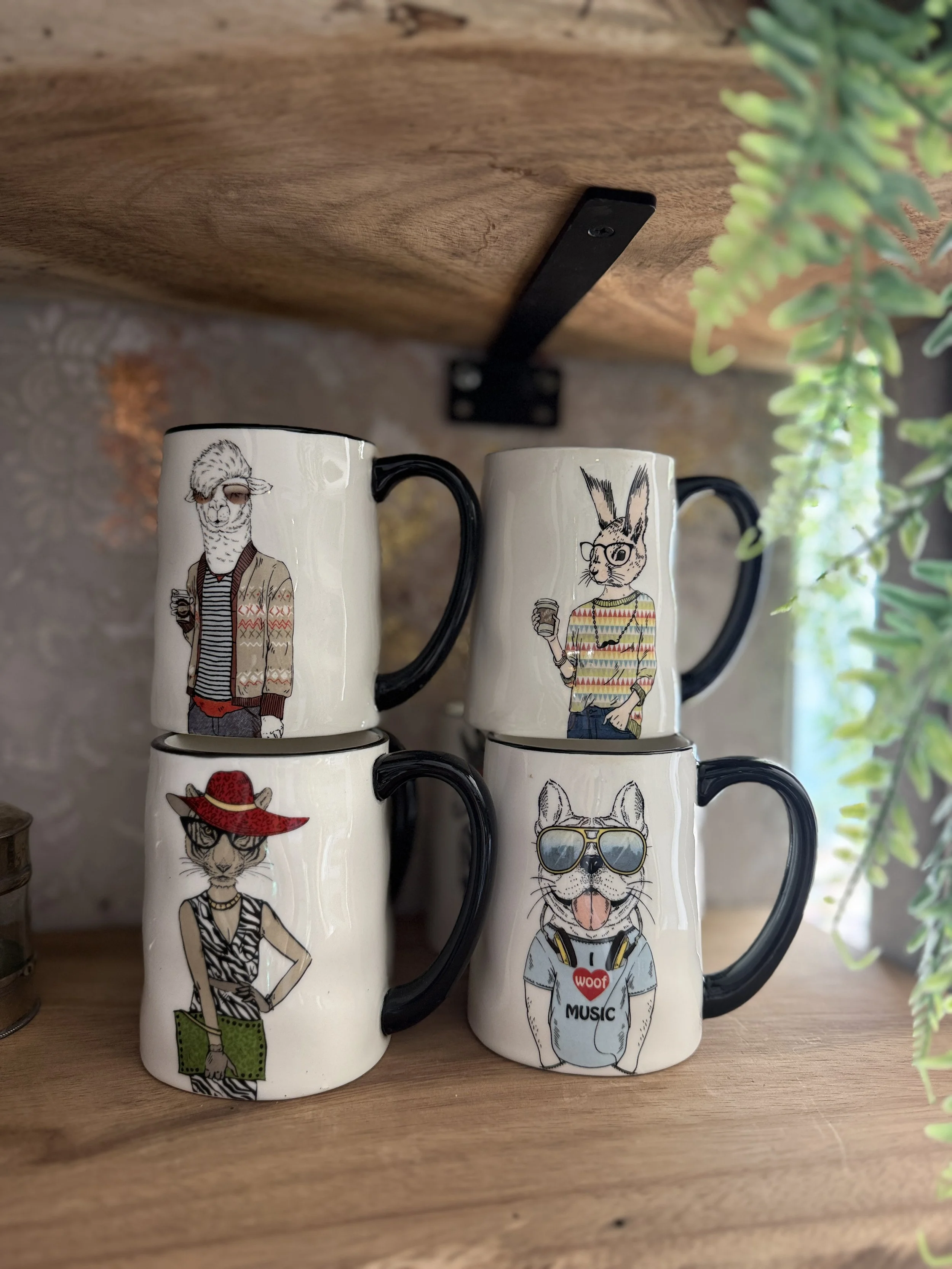 Four ceramic mugs with animal illustrations, stacked two on top of two, on a wooden shelf, with a wooden and brick background and green plants nearby.