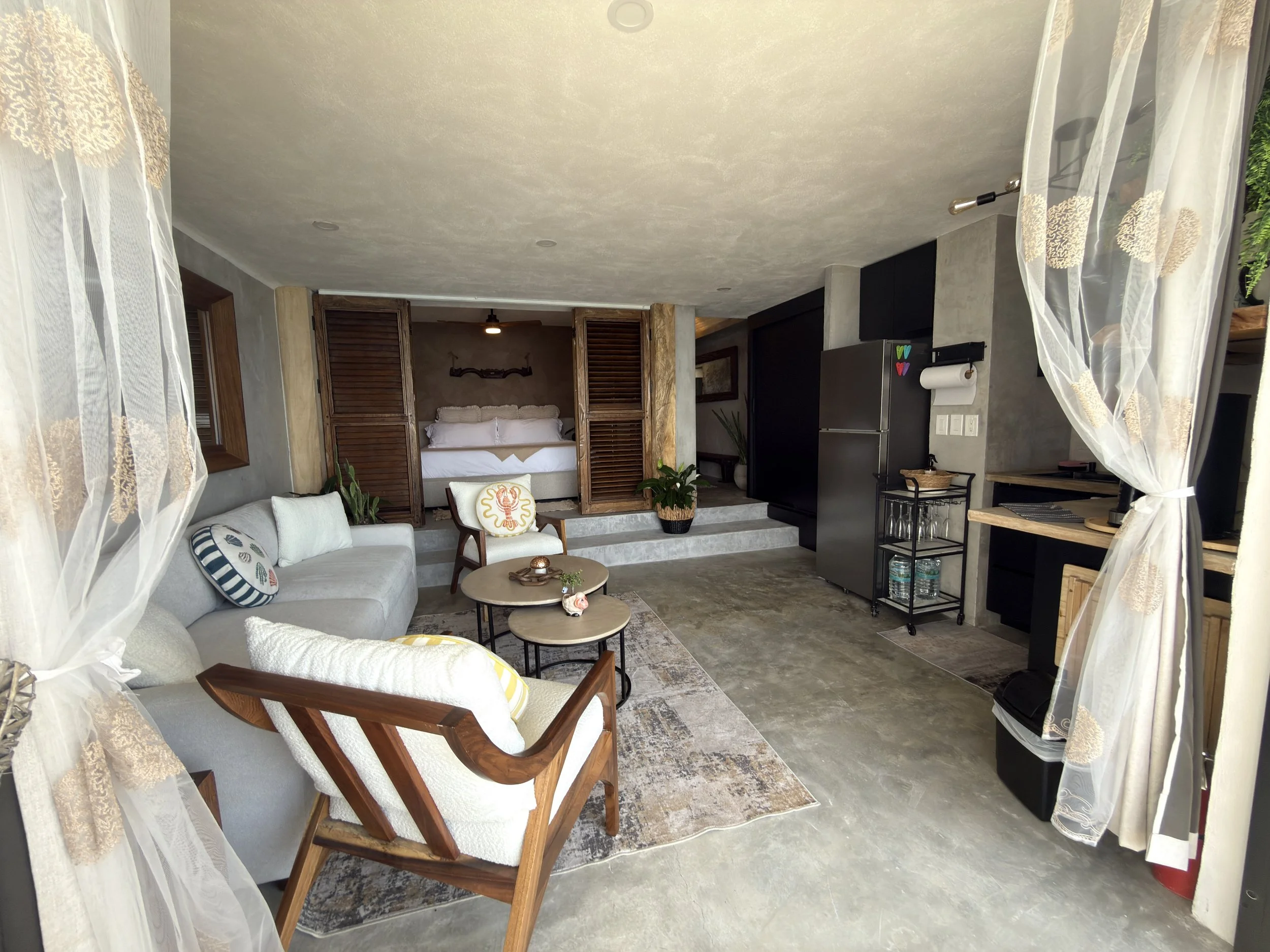 Living room with a sofa, armchair, coffee tables, and a view into a bedroom with a bed behind louvered wooden doors.