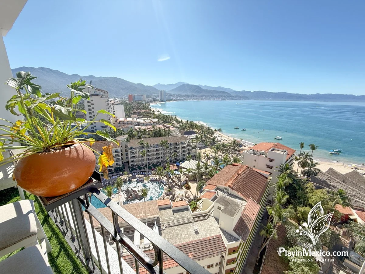 View from a high-rise balcony overlooking a beach, ocean, and distant mountains, with buildings, palm trees, and a swimming pool below.