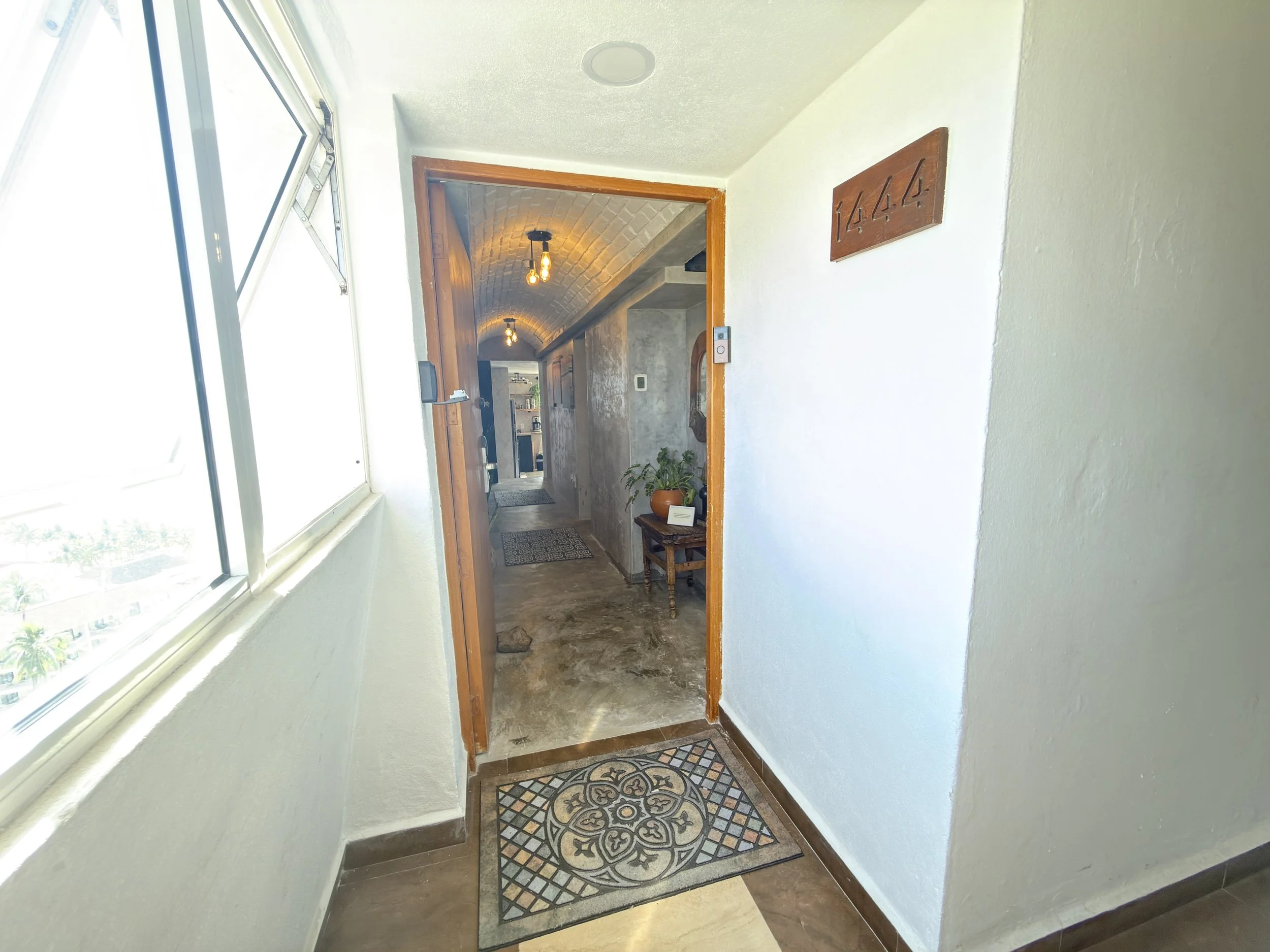 View of a hallway inside a house with white walls, a window on the left, and a brown door frame leading to a corridor with exposed brick ceiling and hanging light bulbs. There is a wooden sign with the number 1444 on the wall to the right.
