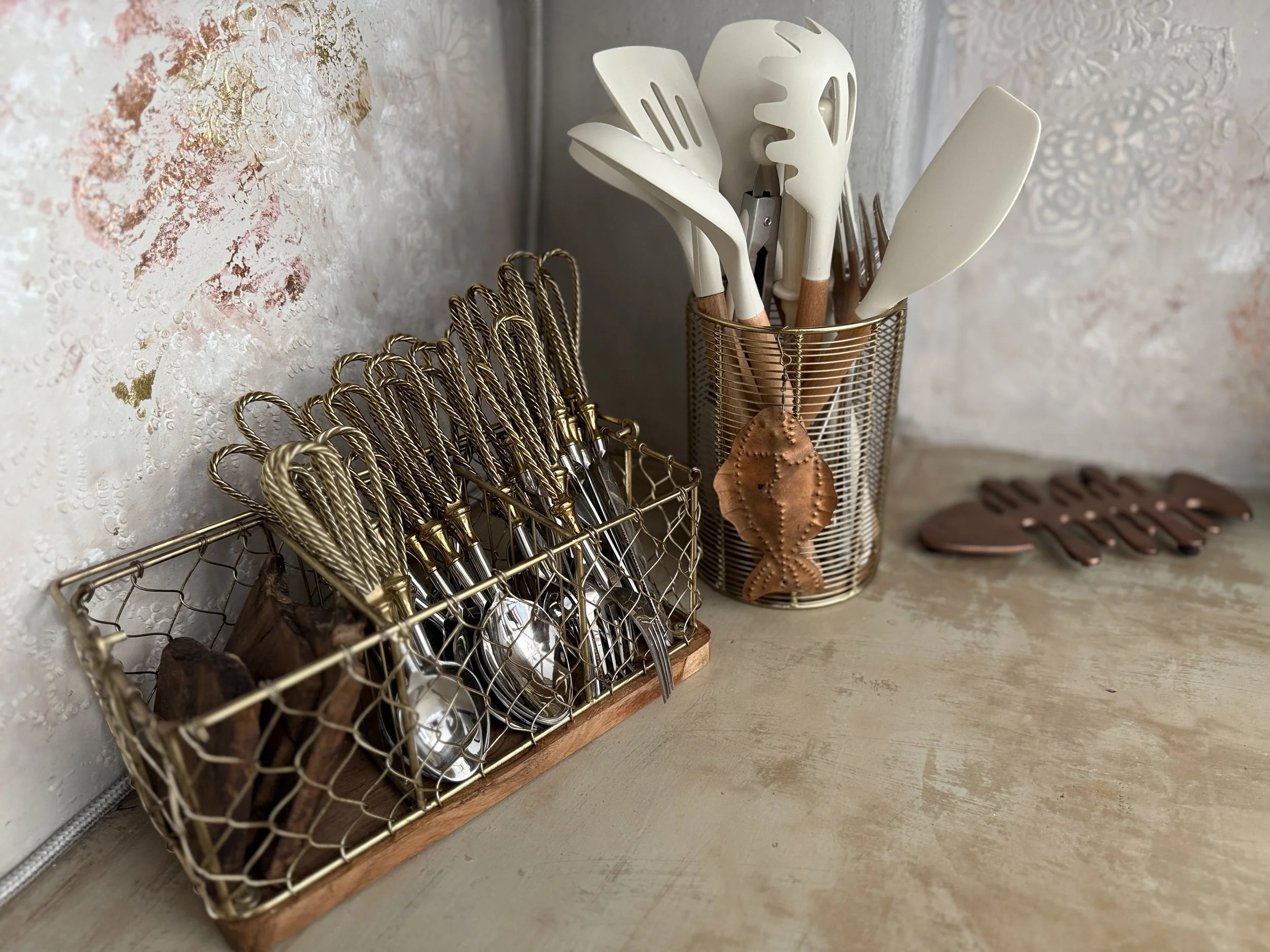 Kitchen utensils on a countertop including a wire utensil holder with white spatulas and forks, a wire spoon holder with copper-colored spoons, and a wire container with copper-colored fish-shaped decorations, against a wallpapered wall.
