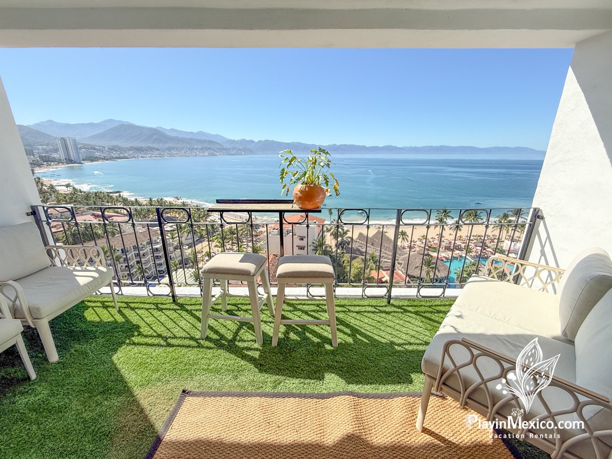 Balcony view over the beach with ocean, mountains, and cityscape in the background. Balcony has outdoor seating, a small table with a potted plant, artificial grass flooring, and a wicker rug.