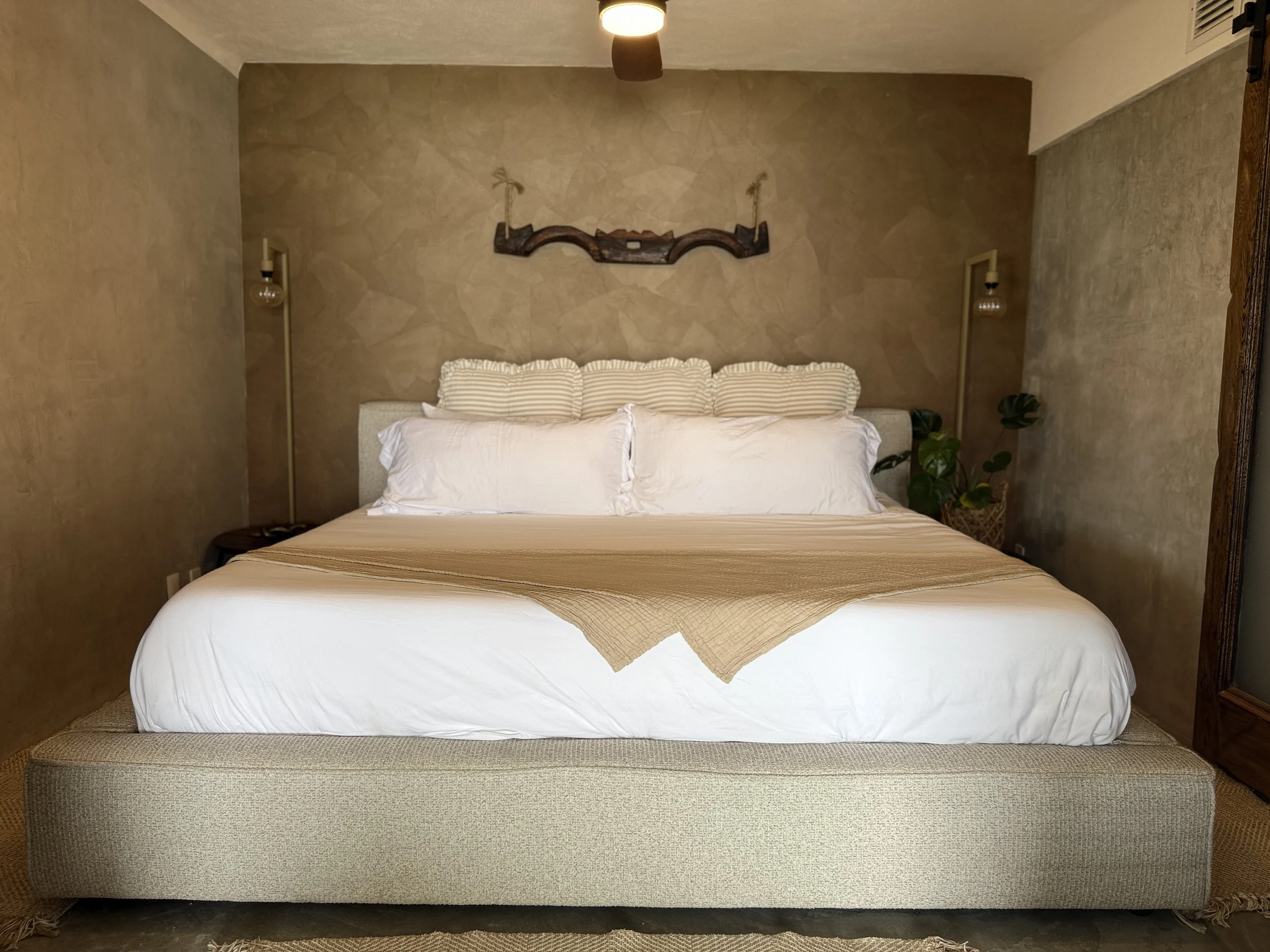 A neatly made bed with a light beige upholstered frame, white pillows and bedding, a beige throw blanket, against a textured brown wall with wall-mounted lamps and a decorative wooden wall piece, near a potted plant.