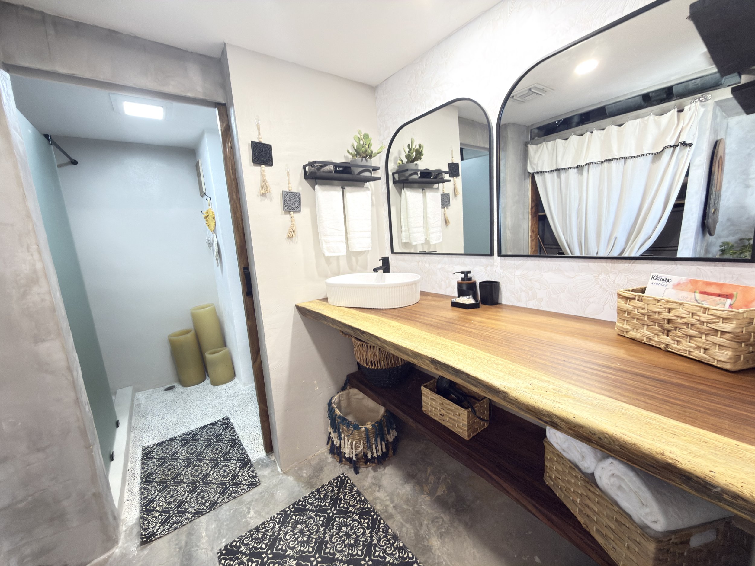 Modern bathroom with a wooden vanity, large mirror, vessel sink, black fixtures, decorative plants, and two black shelving units with towels. There's a small shower area with candles and a textured wall, and a window with white curtains.