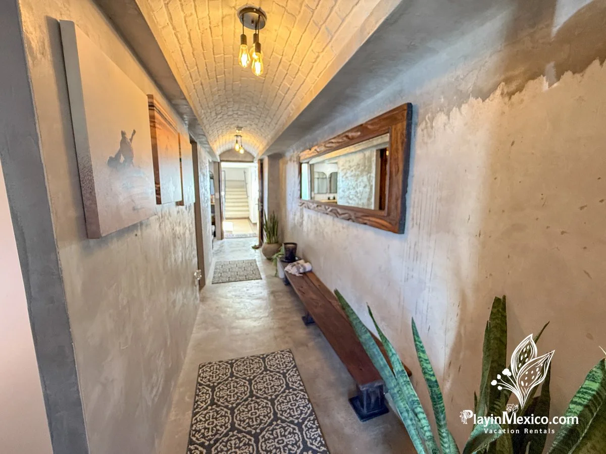 Long hallway with beige textured walls, arched brick ceiling, wooden framed mirror on right wall, paintings on left wall, hanging light bulbs, potted plant and bench at end, patterned rug on concrete floor