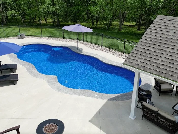 Ground-level view of a Thursday Pools Sun Day 16x39 freeform fiberglass pool in Maya color, featuring a 16-inch decorative coping band and concrete pool deck, with outdoor lounge seating and a fire pit table.