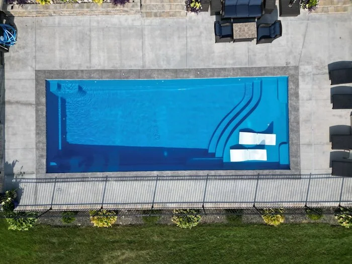 Overhead aerial view of a Thursday Pools Aspen 16x40 fiberglass inground pool in California Blue with tanning ledge chairs, 16-inch decorative coping, and a concrete patio. 