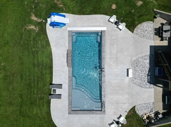 Aerial view of a Thursday Pools Aspen 16x40 fiberglass inground pool in Diamond with a sheer descent waterfall, automatic safety cover, artisan handrail, and concrete patio with rock landscaping.