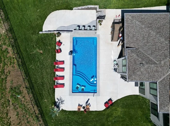 High aerial view of a complete Kansas City backyard pool and patio by Tiger Pool and Patio—Thursday Pools Aspen 16x35 in California Blue with a Grand Effects stainless steel waterfall, sunken outdoor kitchen, and expansive concrete entertaining area.