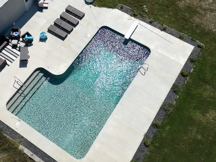 Angled aerial view of a 16x40x16x26 L-shaped diver vinyl liner pool with GLI Sedona liner and sand gunite, featuring a concrete pool deck with lounge seating beside a rural Kansas City outbuilding.
