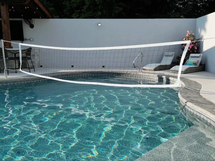 A Tiger Pool and Patio 20x42 custom lagoon vinyl liner pool with Merlin Beachside Tile liner and installed volleyball kit, showing the net stretched across the pool width with lounge chairs, flowering plants, and a pergola bar area along a white priv