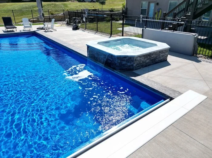 Close-up of a Cardinal fiberglass spillover spa with stone veneer cascading into a Thursday Pools Aspen 16x40 in Maya Blue, a premium pool and spa installation by Tiger Pool and Patio in Kansas City.