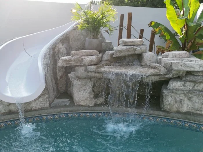 Close-up of the Rico Rock faux rock slide and waterfall built into a 20x42 custom lagoon vinyl liner pool with Merlin Beachside Tile liner, showing the white curved slide, active multi-tier waterfall, and tropical plant accents.