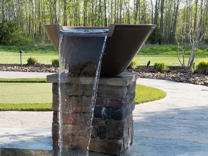  Close-up of a copper water bowl spilling over a stacked stone pedestal beside a custom vinyl liner pool, installed by Tiger Pool and Patio as part of a 20x42 lagoon-style backyard pool design.