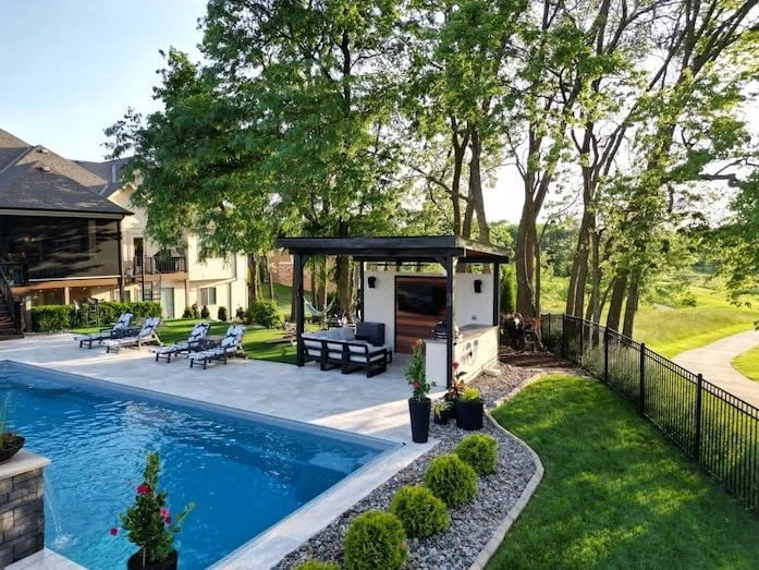A Kansas City backyard pool and patio by Tiger Pool and Patio featuring a Thursday Pools Goliath 16x41 in Diamond with travertine pavers, a modern pergola, an outdoor TV, and a turf lounge area.
