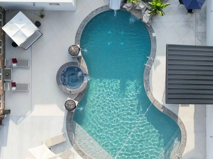 Aerial drone view of a Tiger Pool and Patio 20x42 custom lagoon inground pool with Merlin Beachside Tile and Gold Coast Floor vinyl liner, featuring a round 12-jet spillover spa, two Grand Effects Legacy fire and water bowls in Beechwood, a Rico Rock