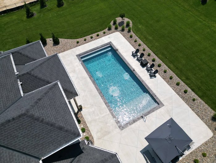 High aerial drone view of a Tiger Pool and Patio 20x40 vinyl liner inground pool with an Island Granite liner, slate coping, and automatic safety cover, set within a large concrete pool deck bordered by curved landscaping beds with ornamental shrubs.