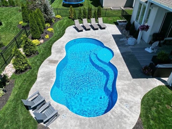 Aerial view of a Thursday Pools Sun Day 15x30 freeform fiberglass inground pool in California Blue with sun deck bubblers, a curved concrete patio, and lush landscaping.