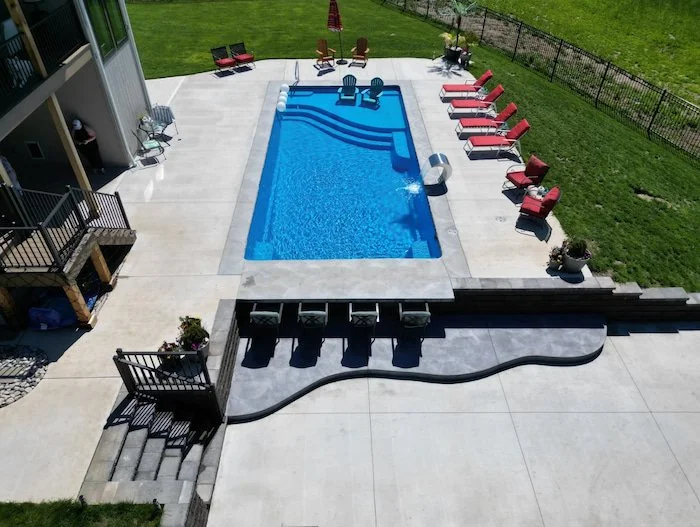 Elevated view of a Thursday Pools Aspen 16x35 fiberglass inground pool in California Blue with a Grand Effects Escala stainless steel waterfall, tanning ledge, and sunken outdoor kitchen with bar seating.